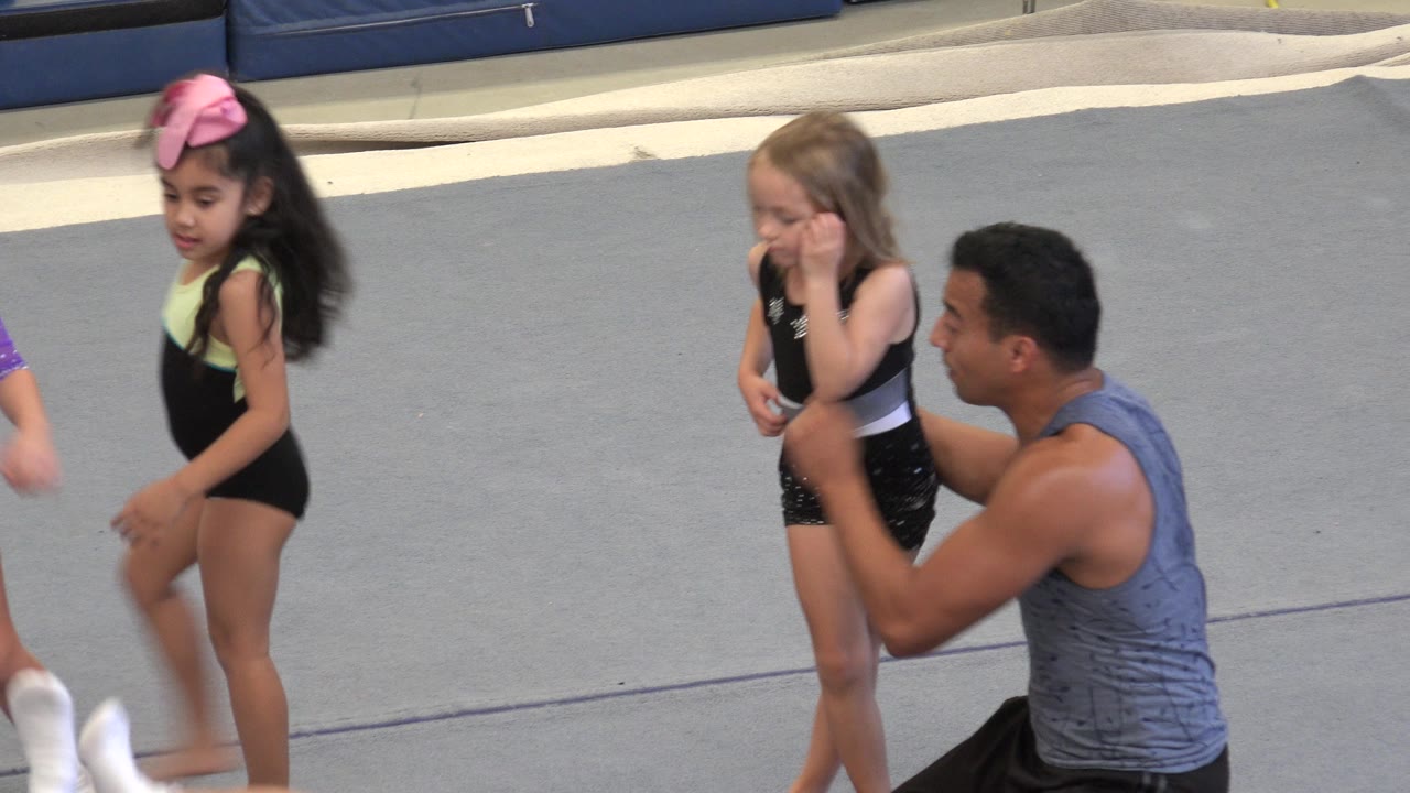 At 0:01 in Cabo San Lucas, the male instructor remains kneeling near a young girl in a black leotard while another child with a pink bow walks across the gray mat.
