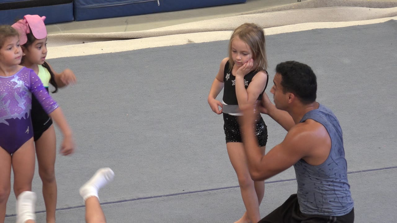 Two young gymnasts stand in front of gray mats as the instructor coaches nearby.