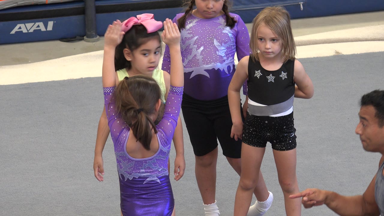 In Cabo San Lucas, Mexico, the young gymnast keeps both arms raised while the kneeling trainer points and three children stand in front of her.