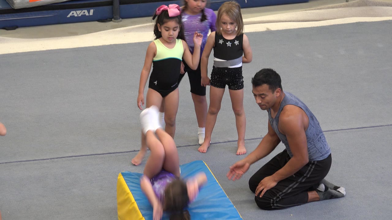 In Cabo San Lucas, Mexico, a young gymnast starts rolling over a blue and yellow wedge mat while three girls stand behind her and an adult coach kneels beside the exercise area.