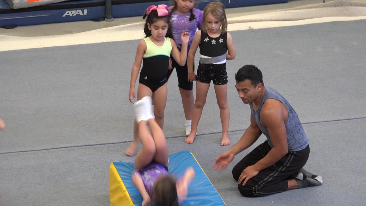 In Cabo San Lucas, Mexico, the gymnast is upside down mid-roll on the wedge mat as the coach kneels at the side and other children wait on the gray gym floor.