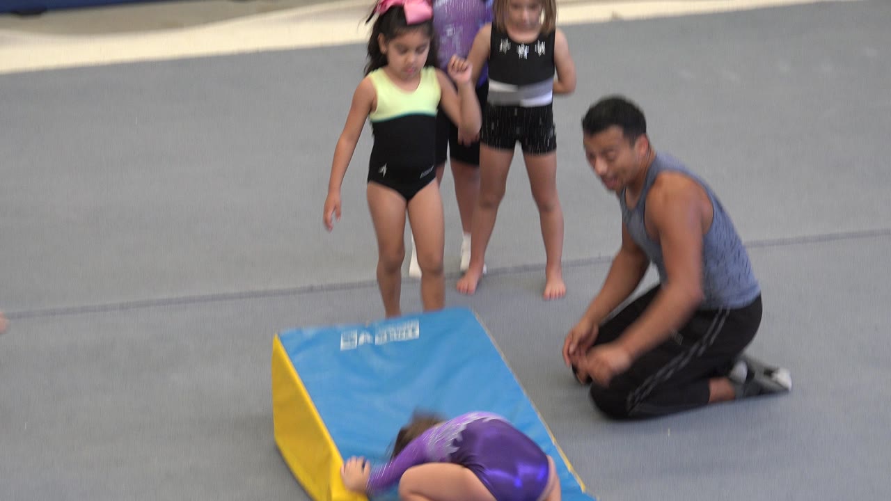 Gymnast finishes rolling off the wedge mat as the coach kneels nearby.