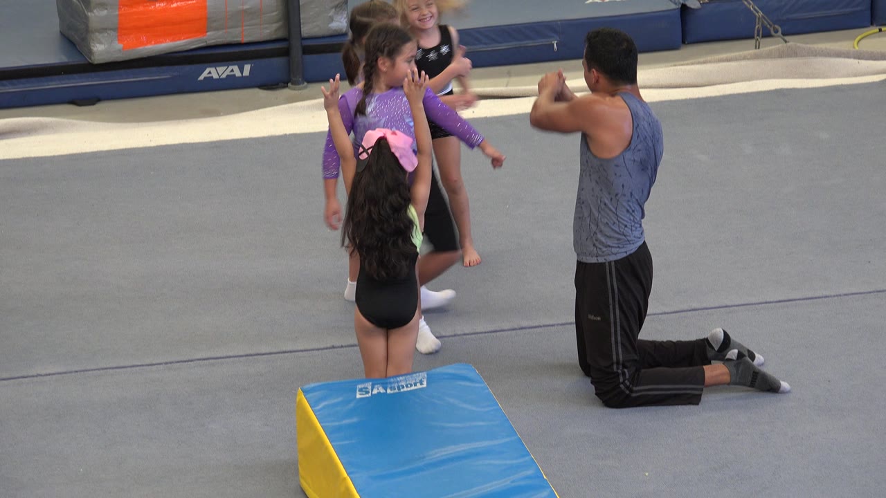 In Cabo San Lucas, Mexico, young girls in leotards stand near a kneeling coach beside a blue and yellow wedge mat on a gray gym floor.