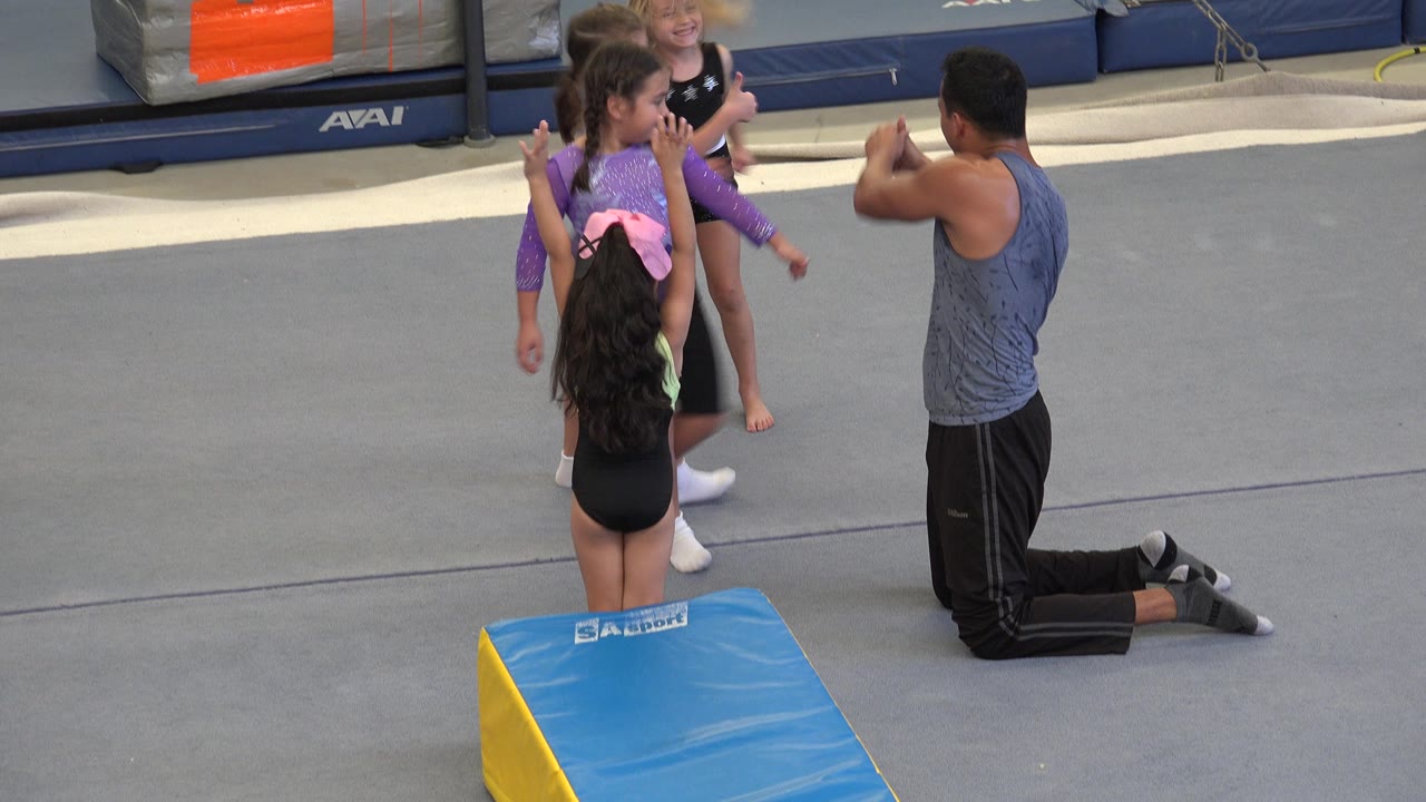 In Cabo San Lucas, Mexico, the girls remain grouped near the blue wedge mat while the kneeling coach gestures toward them during practice.
