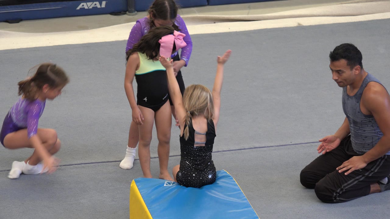Young girls in leotards gather around a blue and yellow wedge mat while an adult coach kneels on the gym floor in Cabo San Lucas, Mexico.