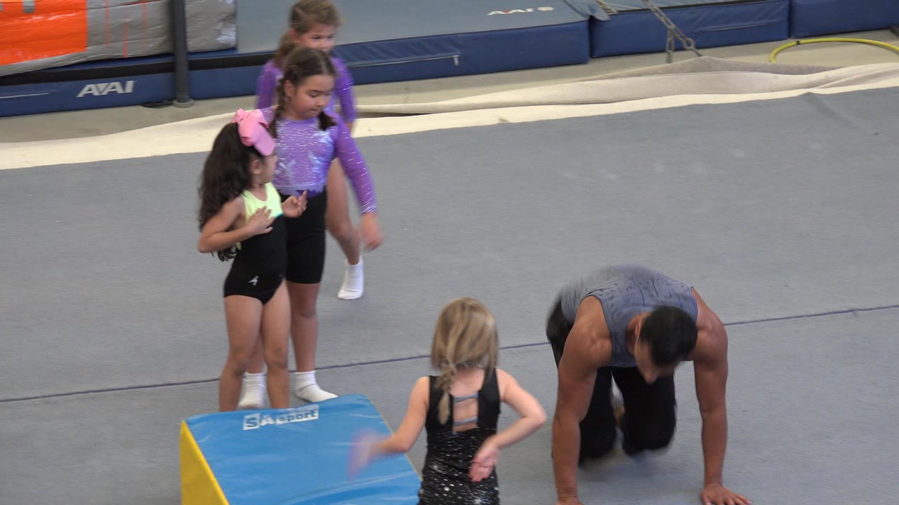 In Cabo San Lucas, Mexico, an adult male instructor crouches on a gray gym floor while four young girls in leotards wait near a blue and yellow padded wedge.