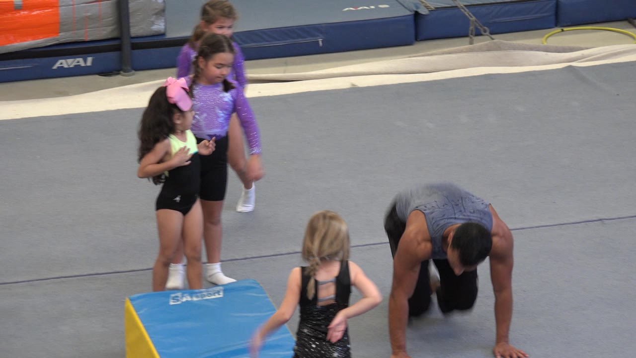 In Cabo San Lucas, Mexico, the instructor is still bent close to the floor while the girls stand around the padded wedge, with the gray gym mat and blue wall padding behind them.