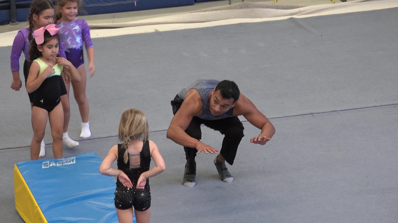 Gymnastics instructor crouches with arms raised as girls watch nearby.