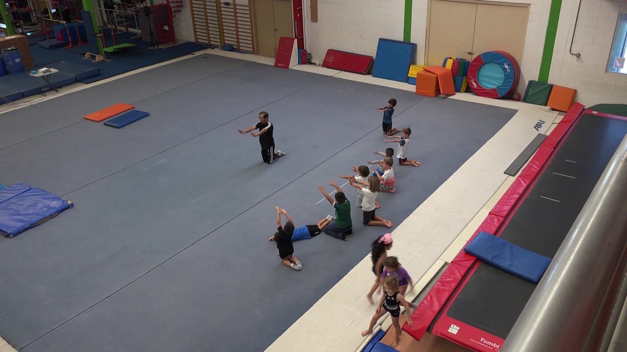 High-angle view in Cabo San Lucas, Mexico, of children sitting on blue gym flooring near an adult instructor, colorful mats, and a trampoline lane along the right side.