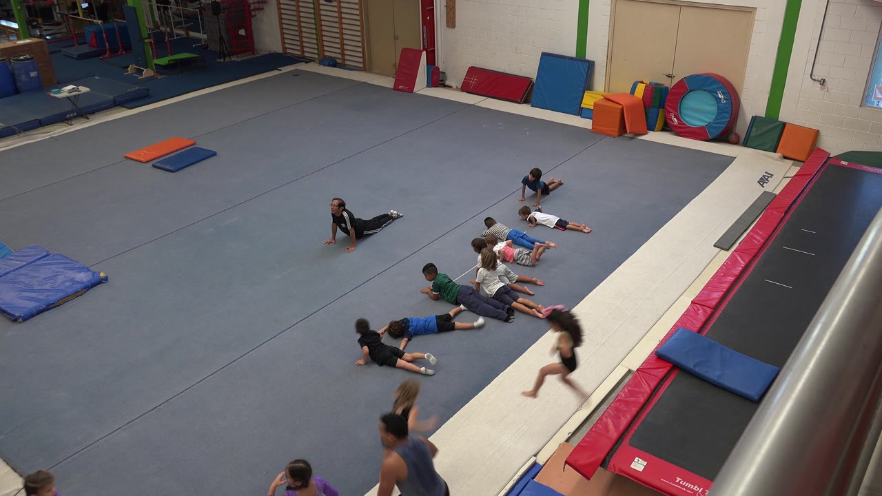 Children lie on a grey padded gym floor in an indoor gymnasium in Cabo San Lucas, Mexico, with a white tumbling lane, red trampoline padding, and blue mats nearby.