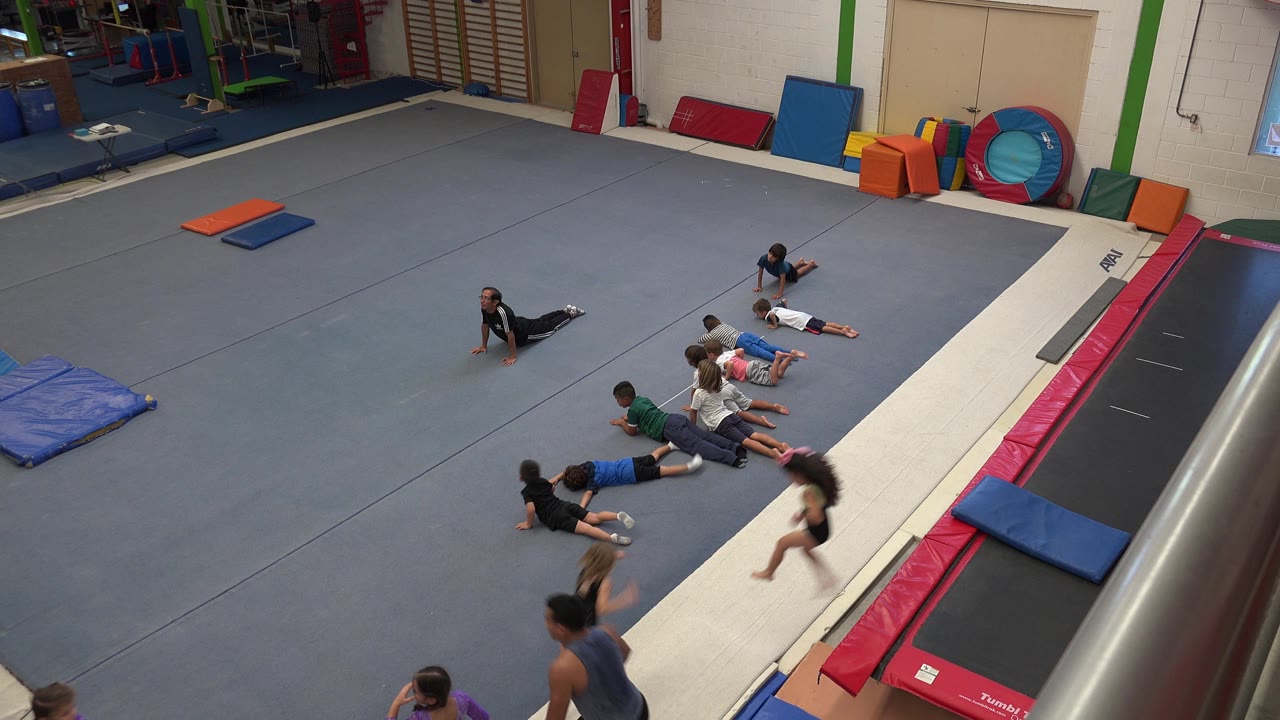 Several children stay low on the grey mat in the Cabo San Lucas gym as a child and an adult or older person move through the lower foreground.
