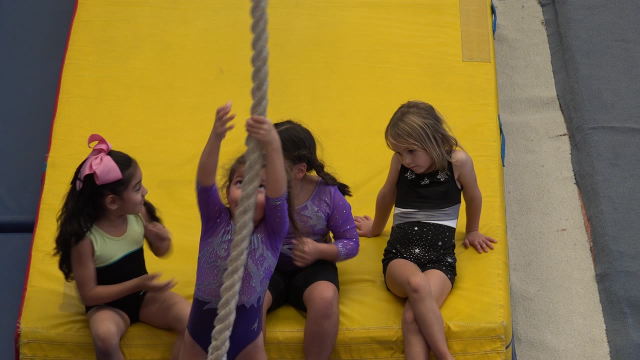 Young girls on yellow mats watch a child holding a rope.