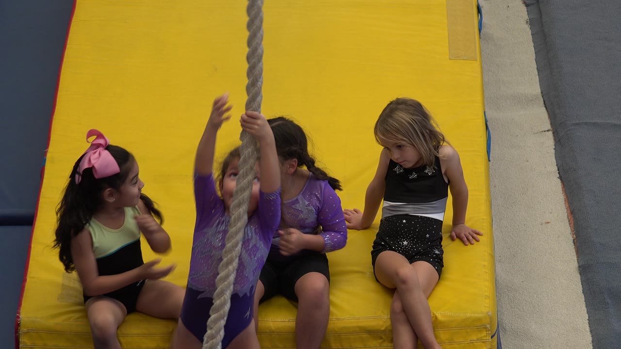 A child in a purple leotard climbs the rope as three girls sit close together on bright yellow mats in Cabo San Lucas, Mexico.