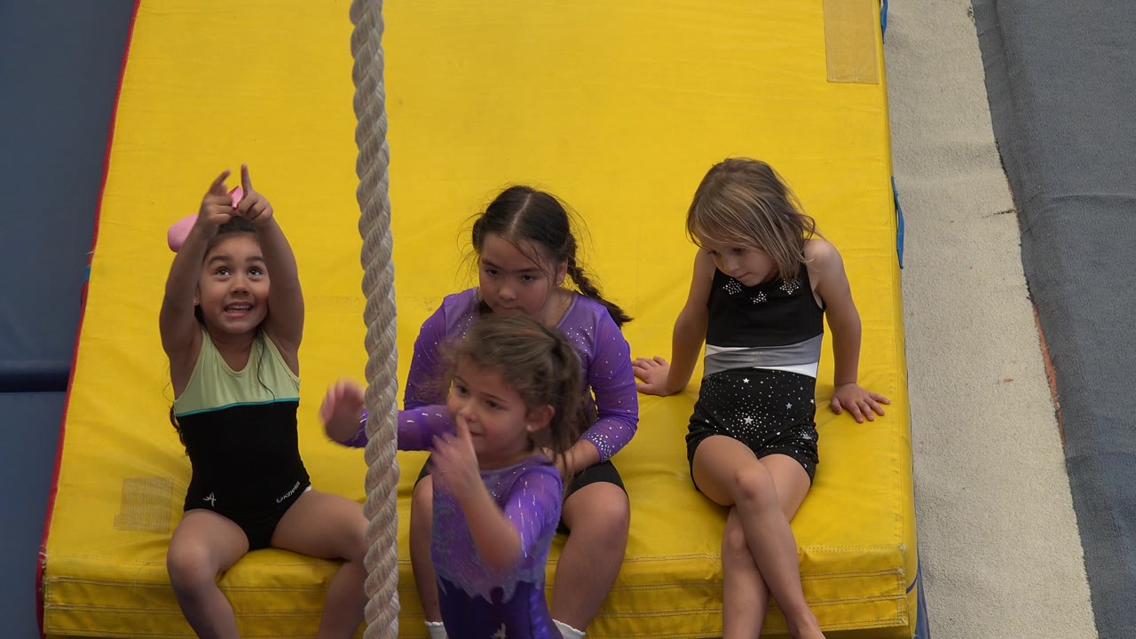 In Cabo San Lucas, Mexico, young children in leotards sit on a bright yellow padded mat as a thick rope hangs in front of them at the start of the clip.