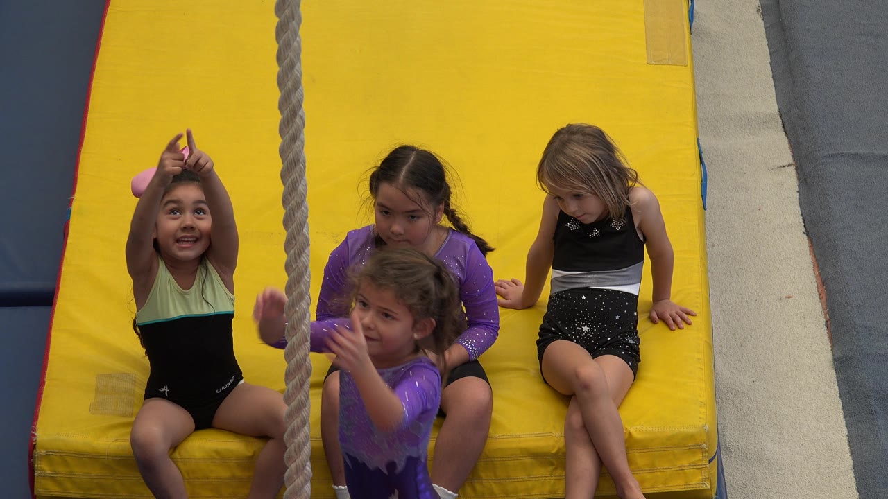In Cabo San Lucas, Mexico, a child smiles and reaches up beside the hanging rope while three other children sit close together on the yellow padded mat.