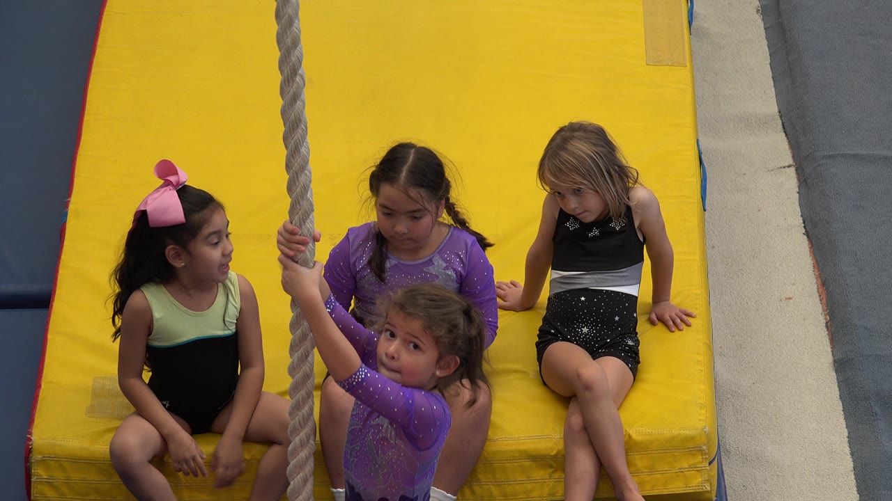 In Cabo San Lucas, Mexico, a child with a pink bow sits near the vertical rope as two children in purple and one child in black watch from the yellow mat.