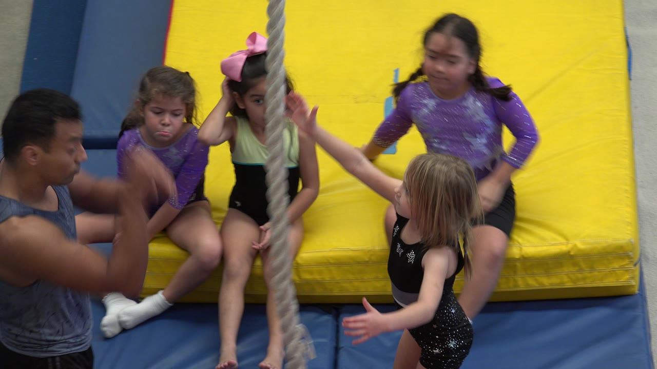 Young girls on padded mats near a climbing rope and adult mentor.