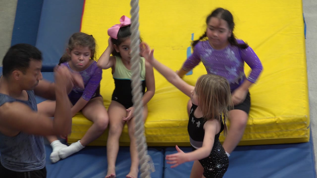 In Cabo San Lucas, Mexico, three girls sit on the yellow mat while another child stands near the rope and the adult mentor gestures from the left edge of frame.