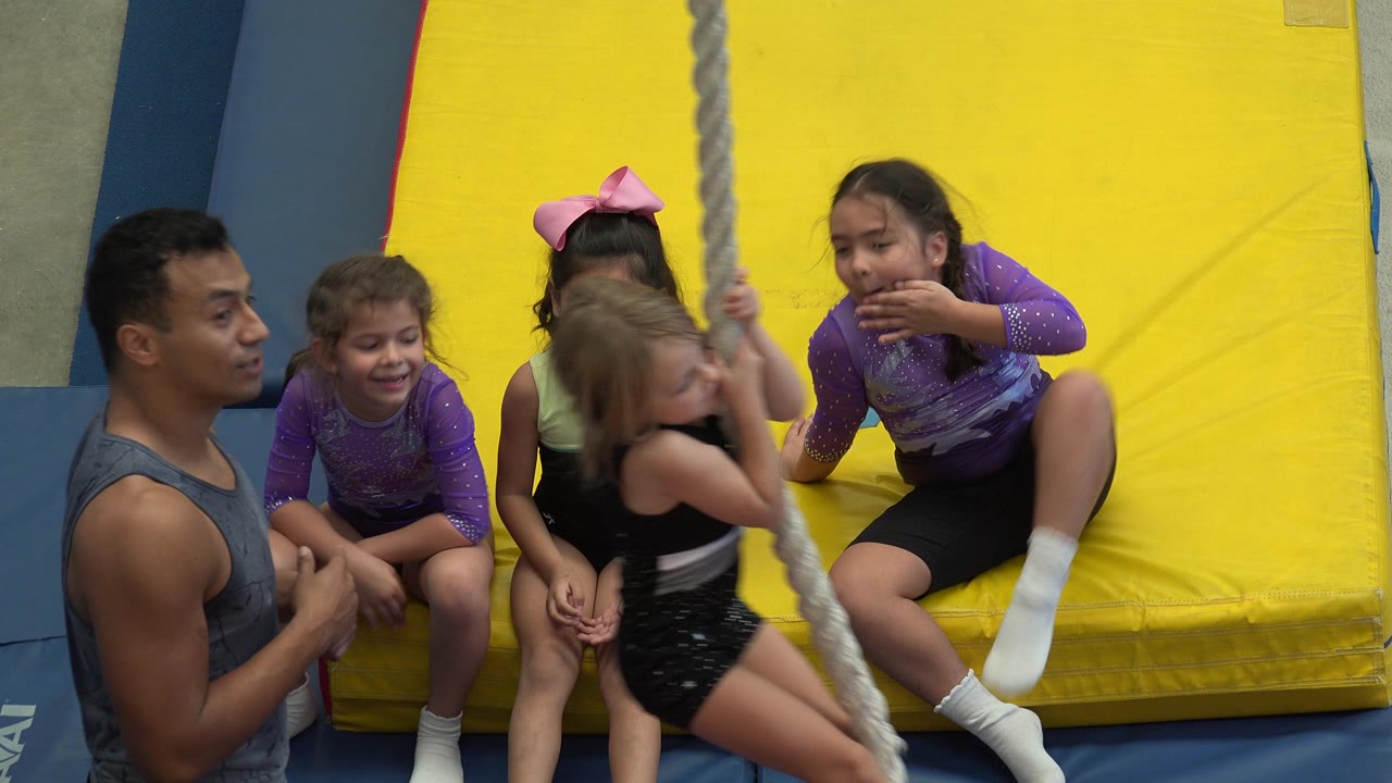 Instructor and four young girls beside a rope on yellow gym mats.