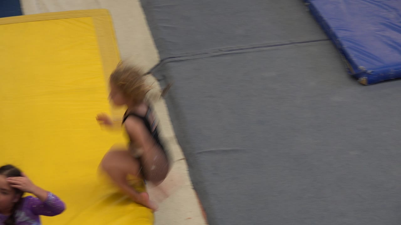 In Cabo San Lucas, a child in a black leotard crouches on the yellow mat, with another child partly visible near the lower edge and gray padding in the background.