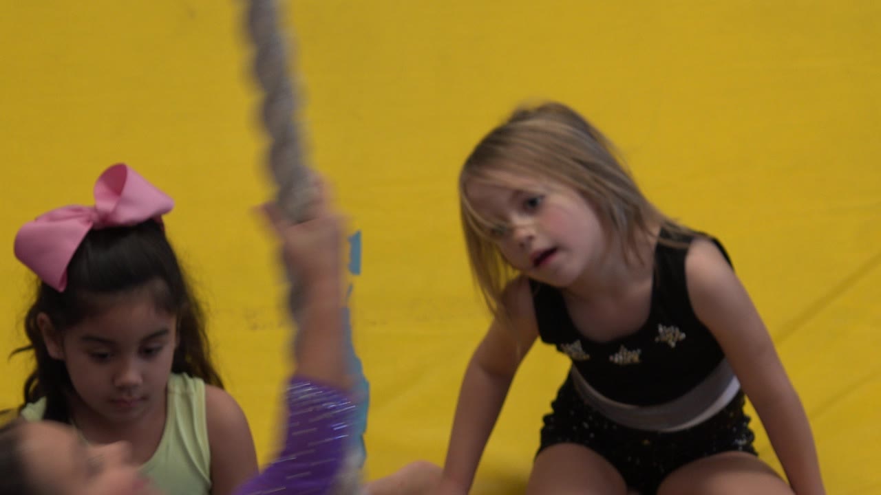 Young girls in gym outfits sit close to a climbing rope against a yellow padded wall in Cabo San Lucas, Mexico.