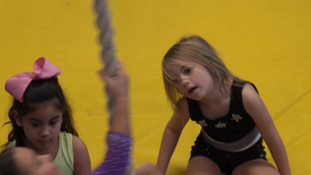 A blonde child in a black gym outfit leans forward near the rope while another girl with a pink bow waits beside her in Cabo San Lucas, Mexico.