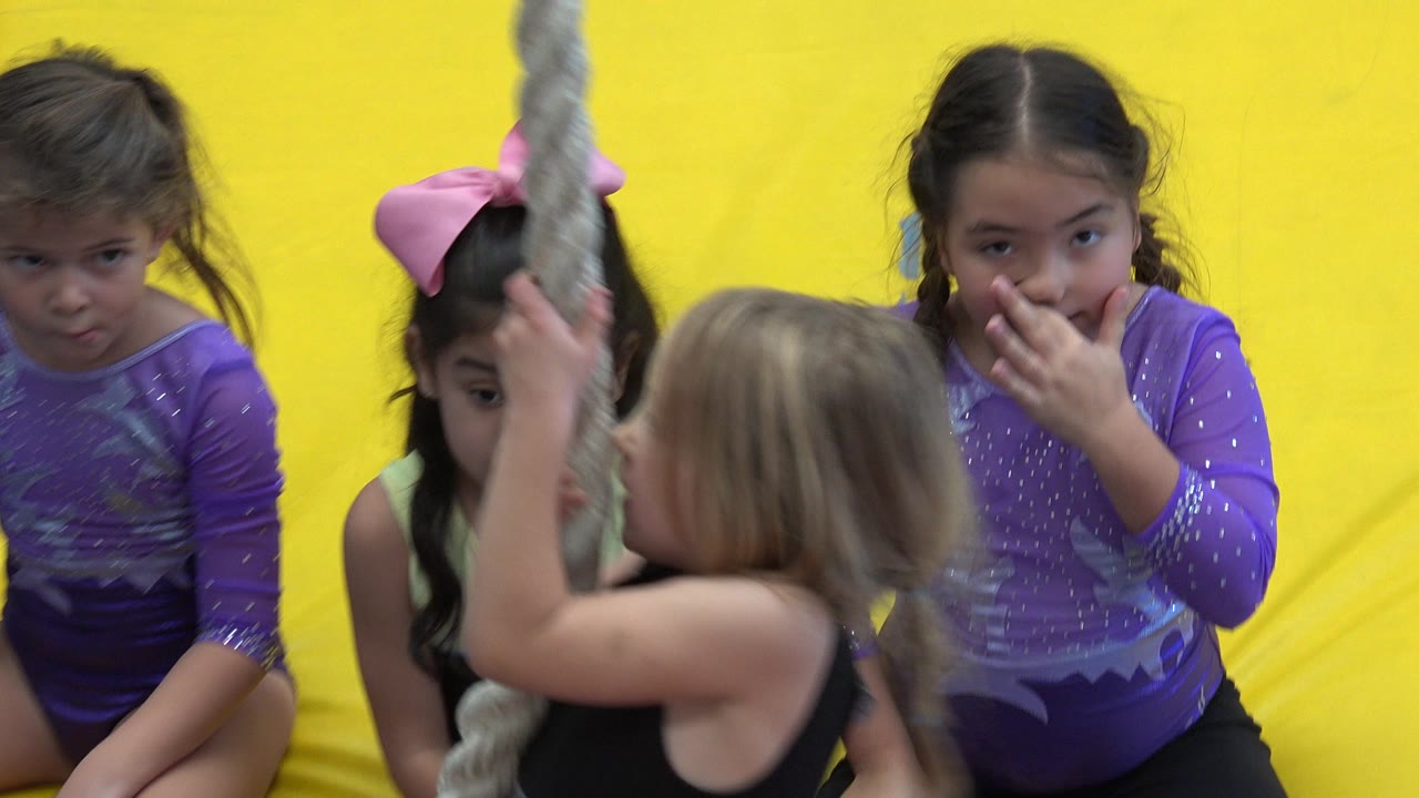 Girls seated near a hanging gym rope against yellow mats.