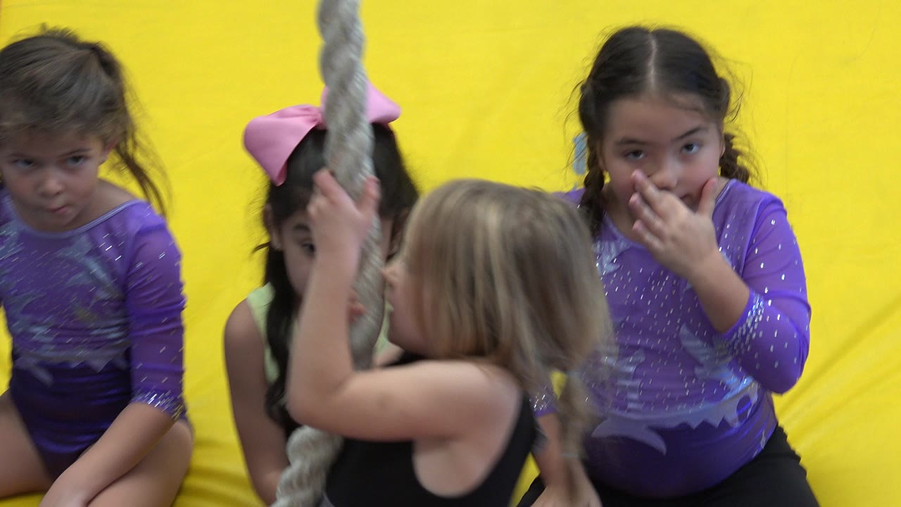 The children remain clustered by the hanging rope in the Cabo San Lucas gym class, with the foreground girl turned toward the rope and two girls facing the camera.