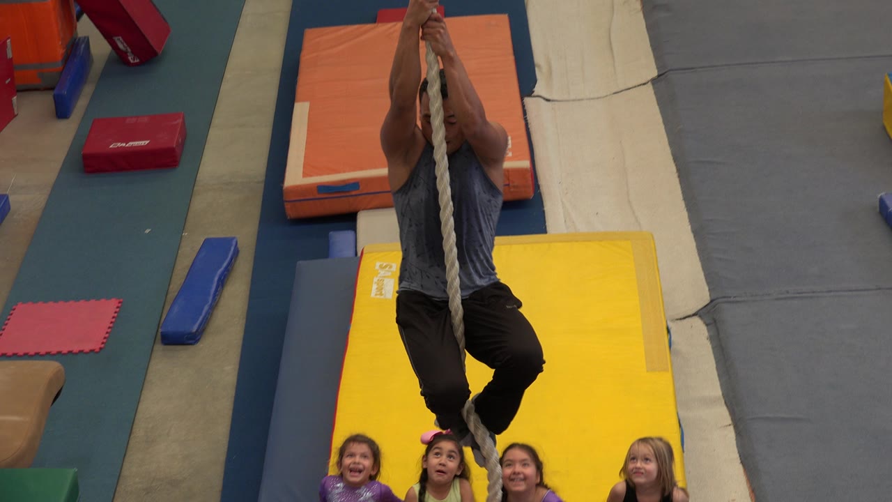 Man holding a rope above gym mats as children laugh below.