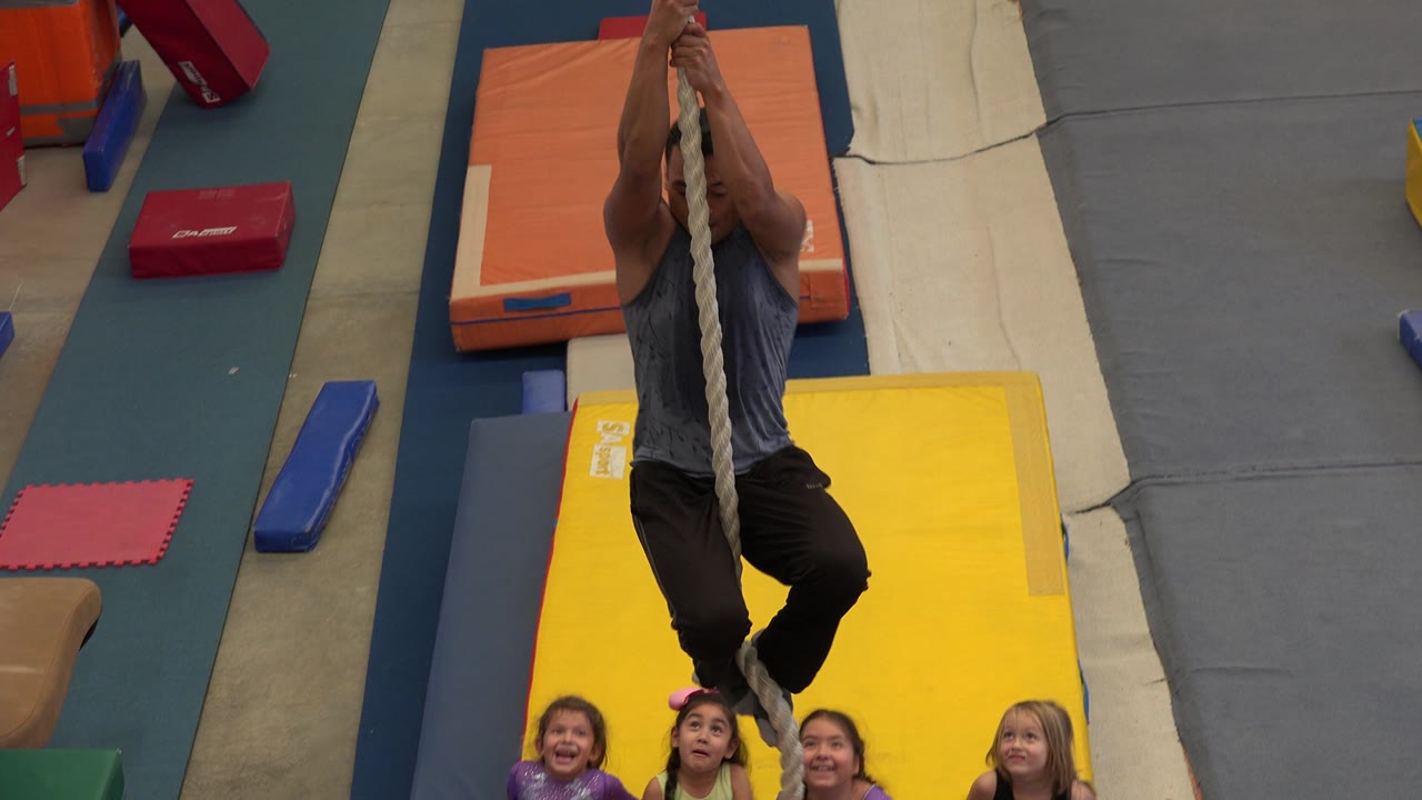 The man remains centered on the rope over the yellow mat, with several children visible along the lower edge in the Cabo San Lucas activity space.