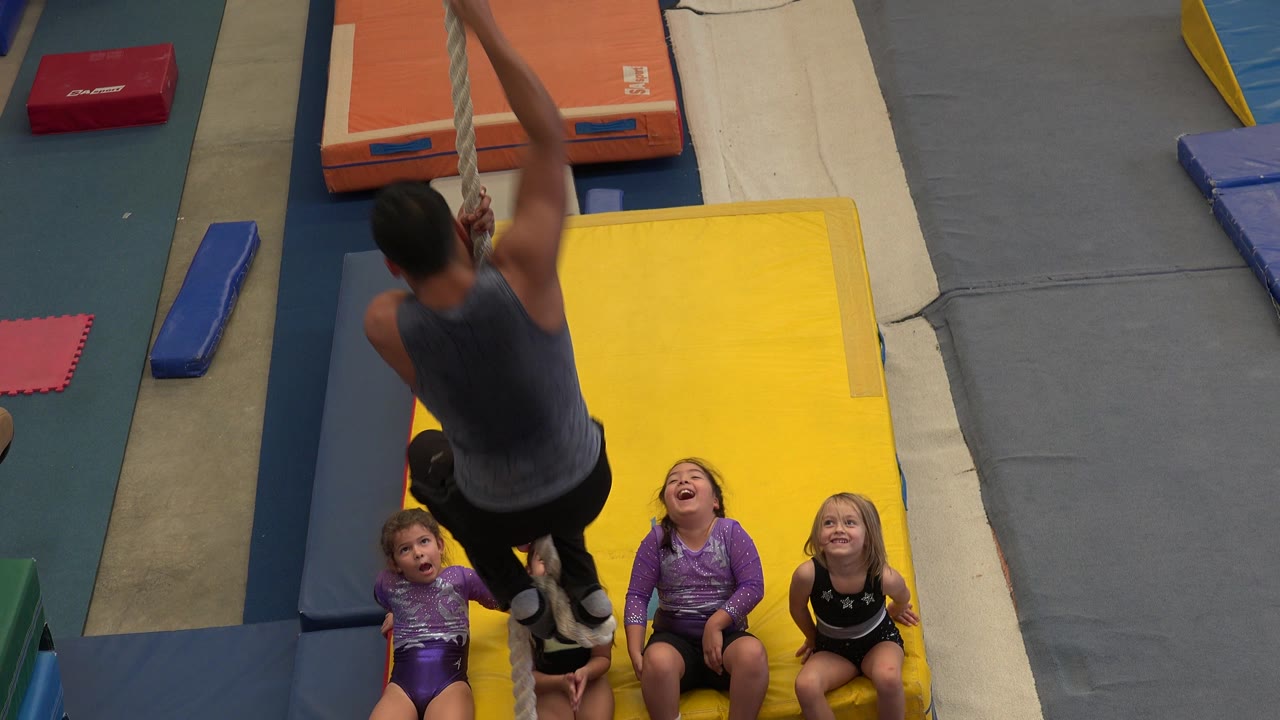 In Cabo San Lucas, Mexico, a man climbs a tan rope while young girls sit below on a yellow mat in an indoor gym area.