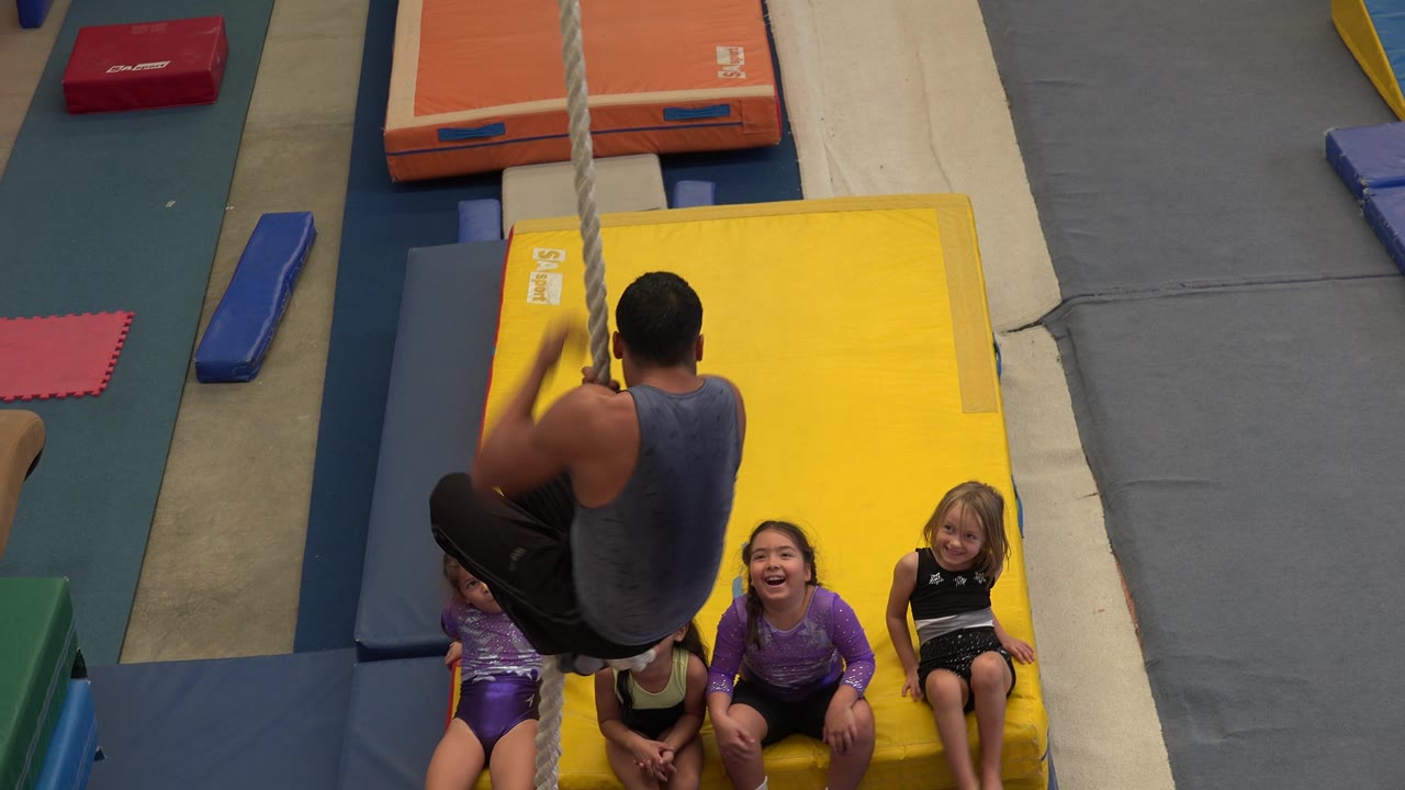 Man holds the rope with knees bent while children sit on the yellow mat.