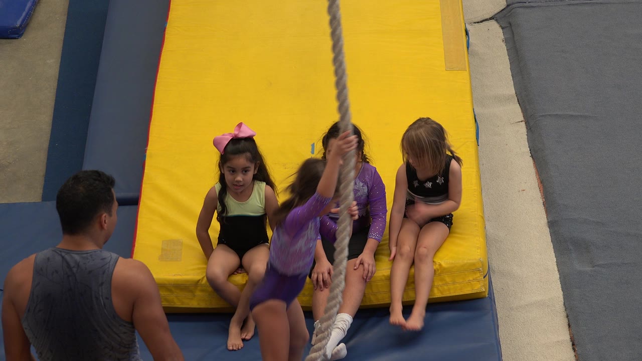 In Cabo San Lucas, young girls sit on a yellow gymnastics mat while a coach faces them and a rope hangs in the foreground.