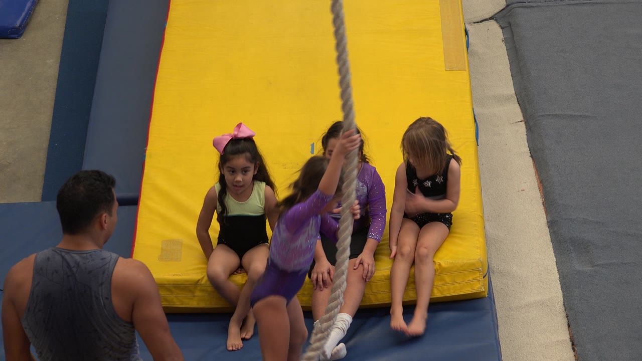 A child moves around the hanging rope as other girls sit on the yellow mat and the coach watches from the lower left.