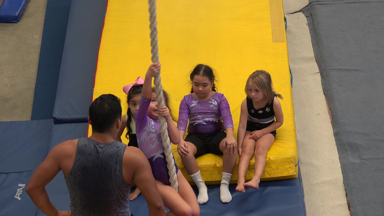 Children sit on a yellow gym mat while a coach stands by a climbing rope.