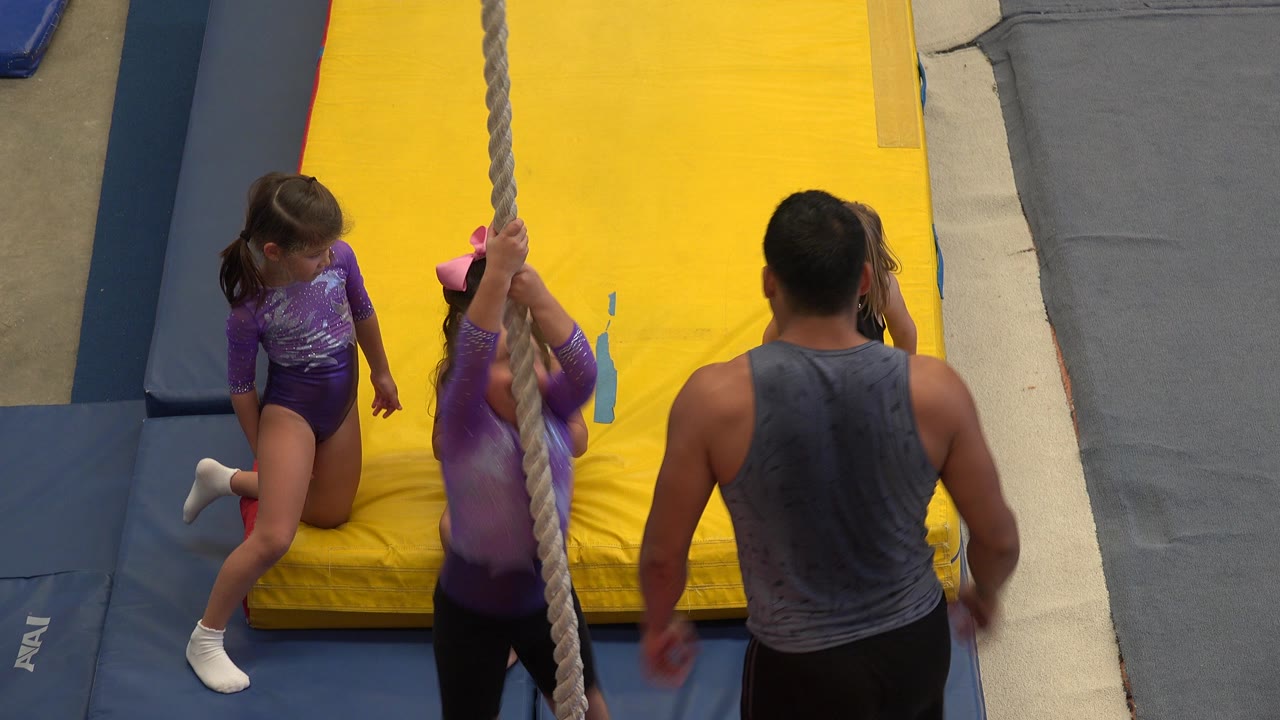 At 0:00 in Cabo San Lucas, Mexico, children stand and sit around a central climbing rope while an adult instructor faces them on padded yellow and blue mats.