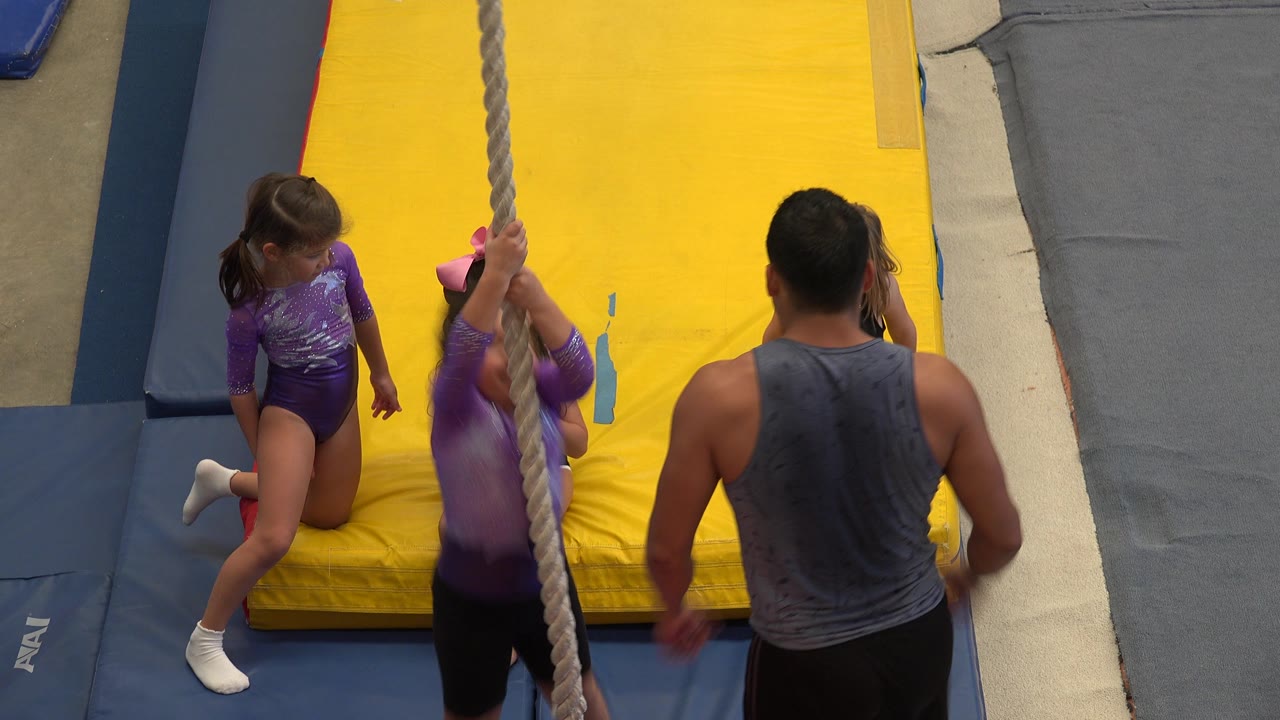 At 0:01 in Cabo San Lucas, Mexico, one child in purple reaches around the rope while another child kneels nearby and the instructor stands close on the padded floor.