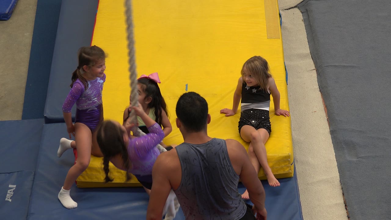 At 0:00, young gymnasts and an adult coach gather beside a hanging rope on blue and yellow padded mats in Cabo San Lucas, Mexico.