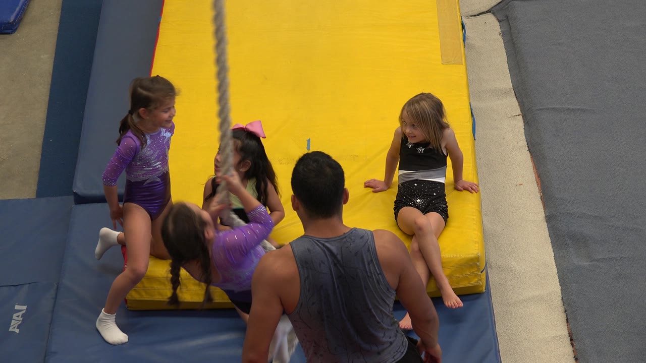At 0:01, the coach stands with his back to camera as children in gymnastics outfits sit and kneel near the rope on the padded floor in Cabo San Lucas, Mexico.