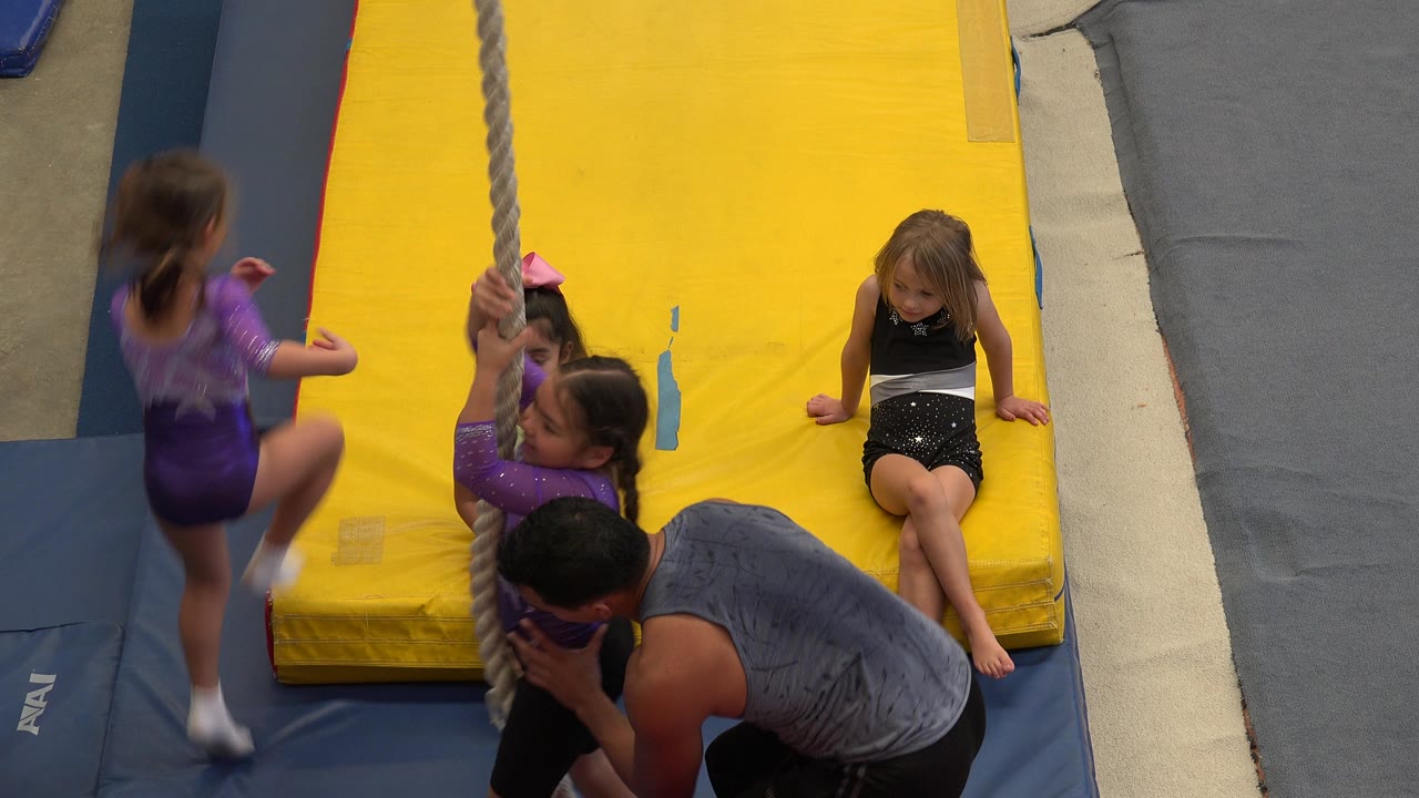 At the start of the Cabo San Lucas clip, young girls and an adult instructor gather around a hanging gym rope on yellow and blue mats.