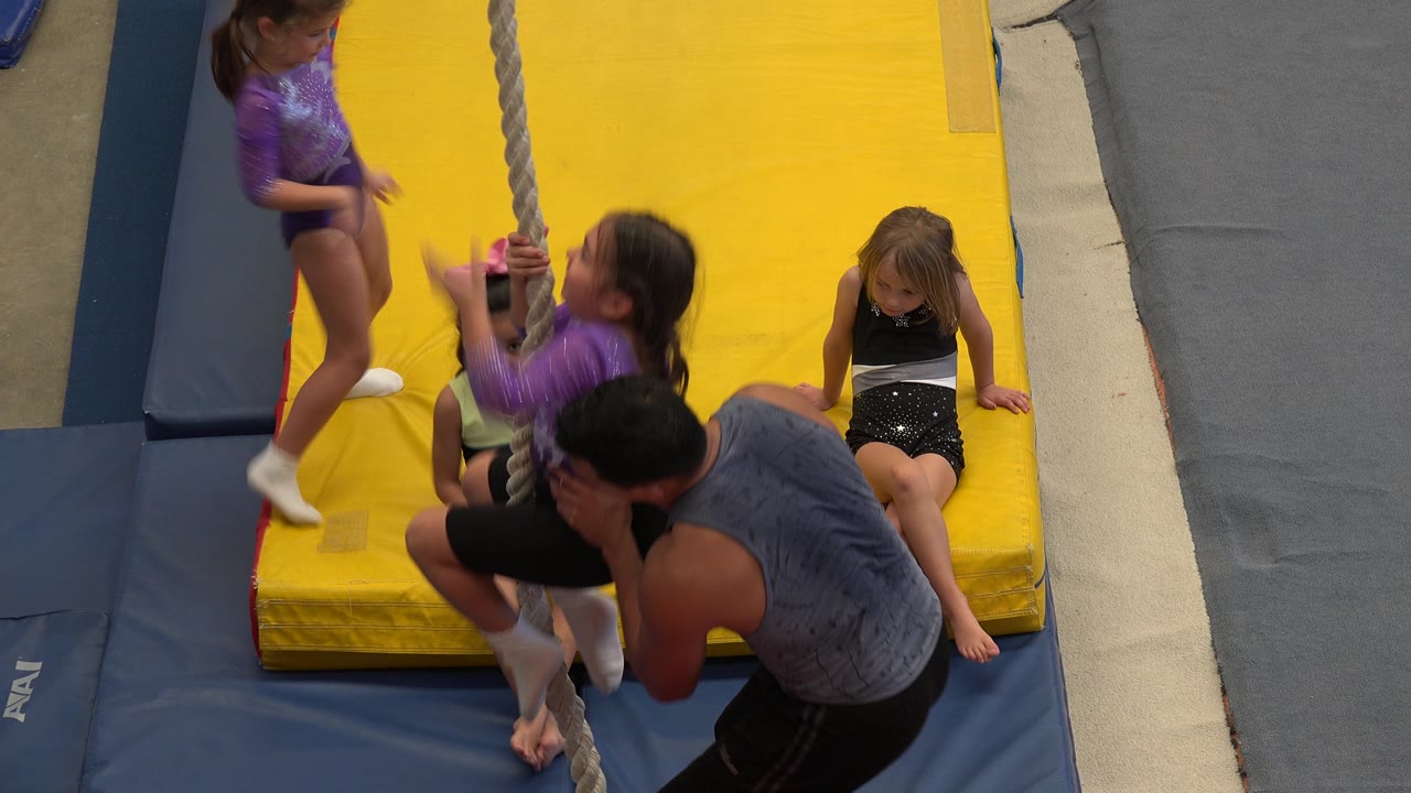 Instructor supports a child on a rope above gym mats.