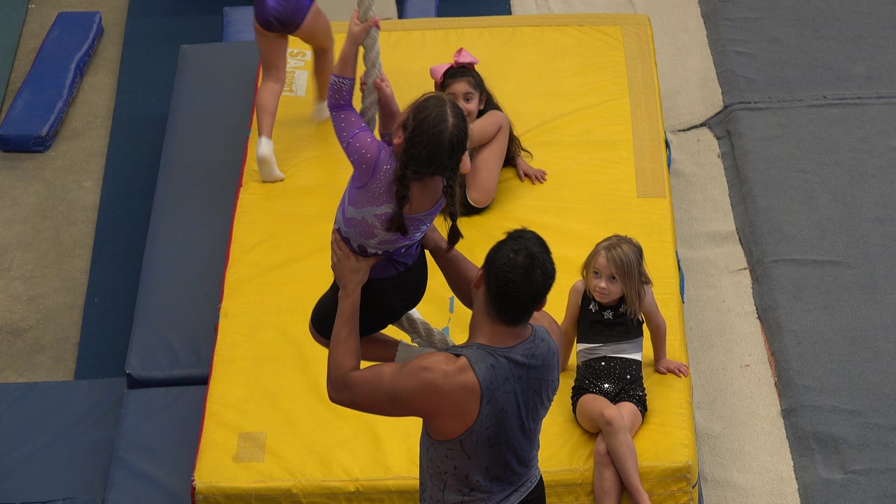 In Cabo San Lucas, Mexico, the child in purple remains lifted on the rope as the adult spotter stands below and a girl with a pink bow watches from the mat.