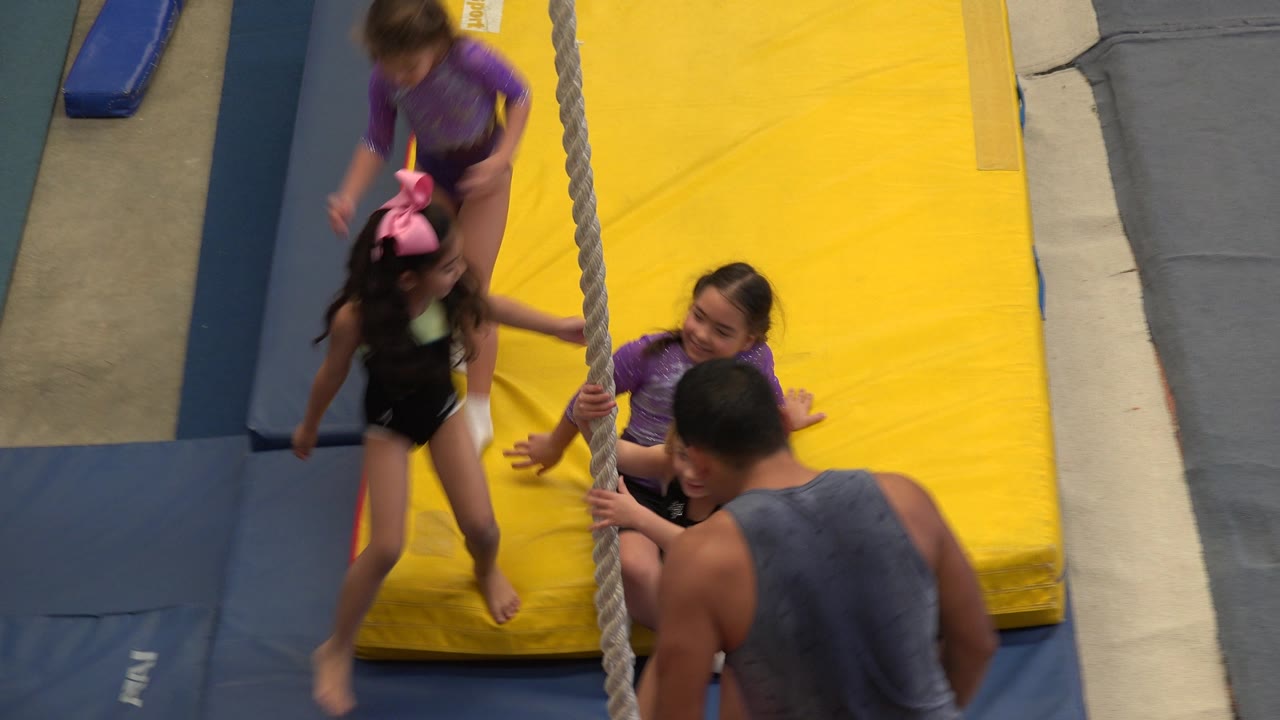 A young girl stands beside the rope as two girls sit near the yellow mat and the adult coach remains in the foreground of the Cabo San Lucas gym scene.