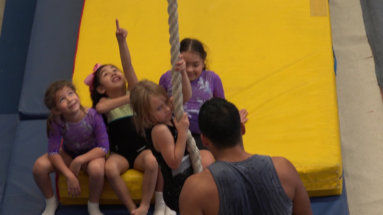 Children sit on gym mats as a blonde girl grips a hanging rope.