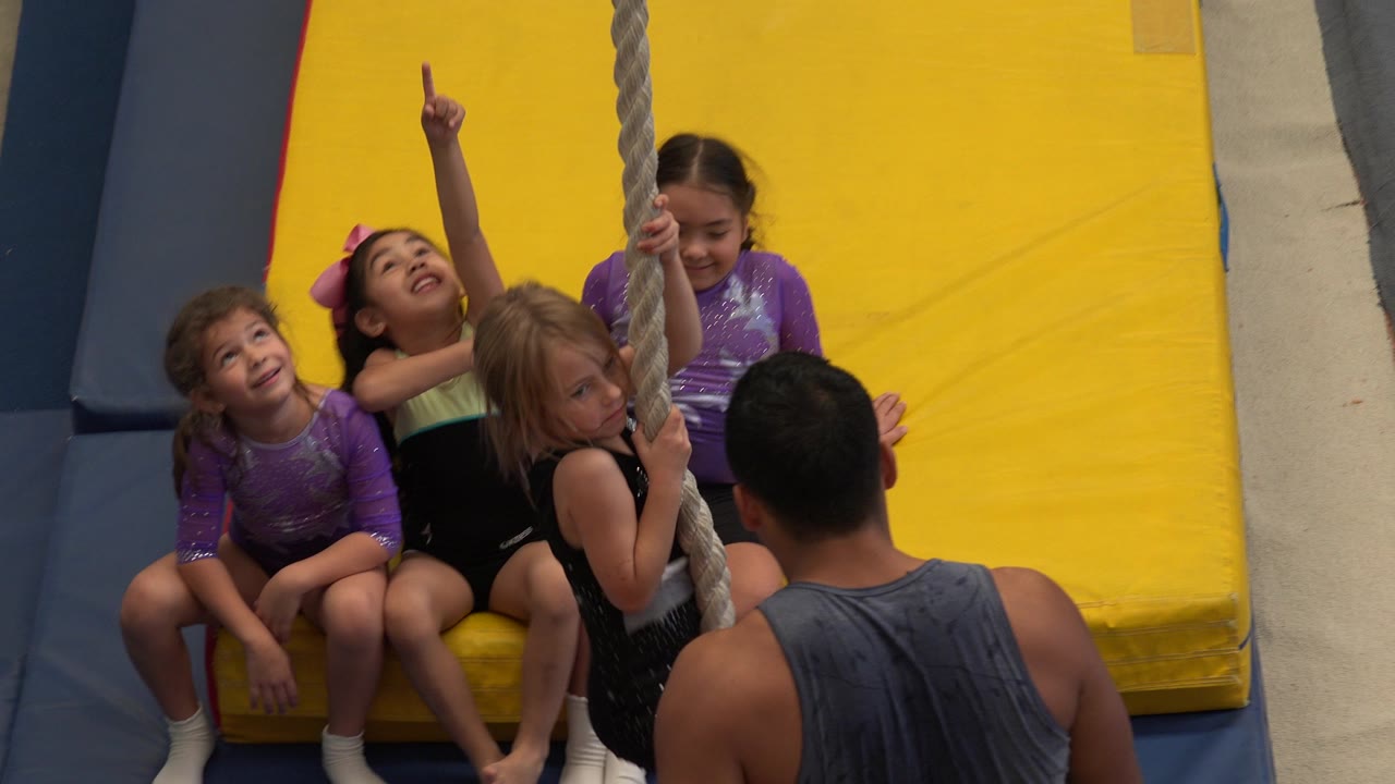 In Cabo San Lucas, one seated girl points upward beside the rope as the blonde child grips it and other children watch from the padded mat area.