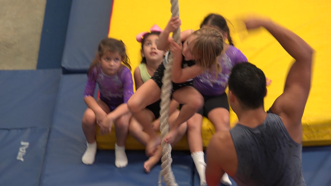 At 0:00, young girls sit on yellow and blue padded mats in Cabo San Lucas while a thick rope hangs in front of them and an adult coach stands close by.