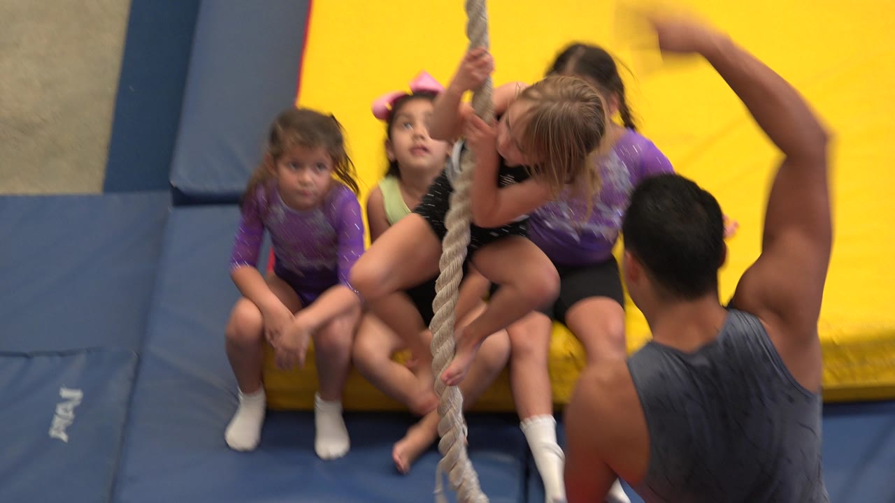 At 0:01, the same group of young girls remains seated on the yellow mat in Cabo San Lucas as the adult coach gestures beside the vertical rope.
