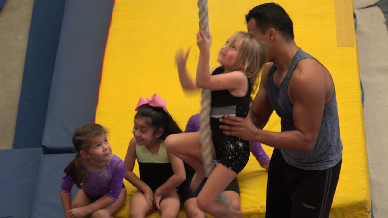 Girl on gym rope with coach and children on yellow mats.