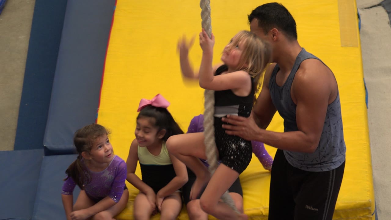 In Cabo San Lucas, Mexico, the girl holds the rope higher as the coach stands close behind her and two young gymnasts look on from the mat.