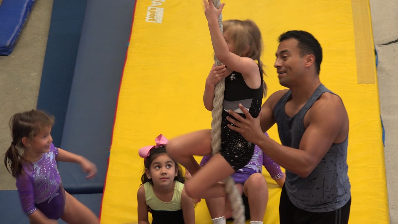 In Cabo San Lucas, Mexico, a coach steadies a young girl gripping a rope while children in leotards gather around yellow and blue padded gym mats.